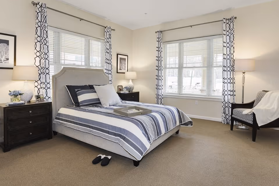 A bright and neatly arranged bedroom with a bed featuring a striped blue and white comforter and multiple pillows. The room has two windows with white blinds and patterned curtains, two dark wooden nightstands with lamps and decorative items, a cushioned armchair with a white throw blanket, and beige carpeting. A pair of slippers is placed on the floor near the bed.