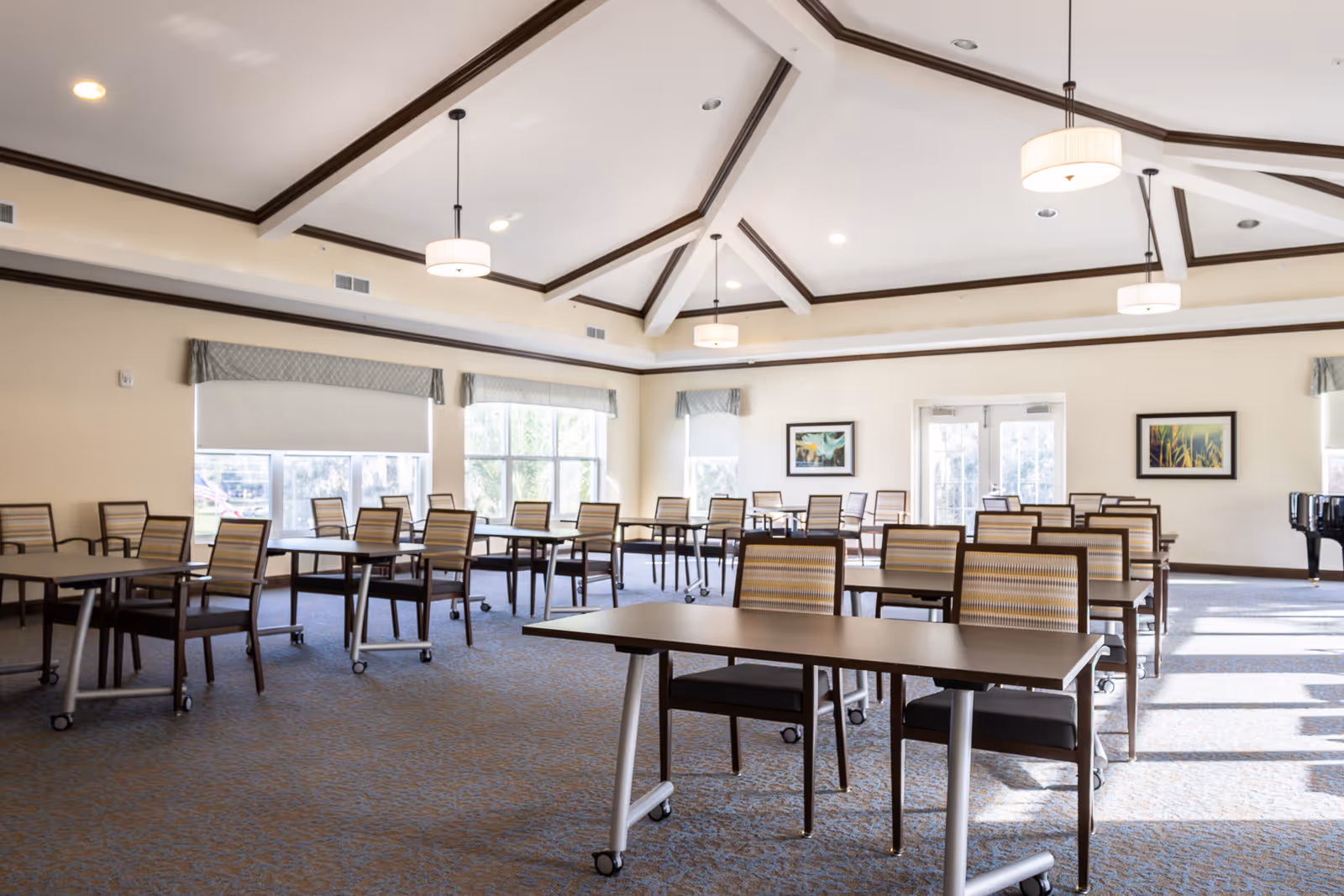 A spacious, well-lit room with multiple tables and chairs arranged in rows, featuring a high vaulted ceiling with exposed beams and hanging light fixtures. Large windows with valances allow natural light to fill the room, and framed artwork decorates the walls. The room has a carpeted floor and double glass doors at the far end.