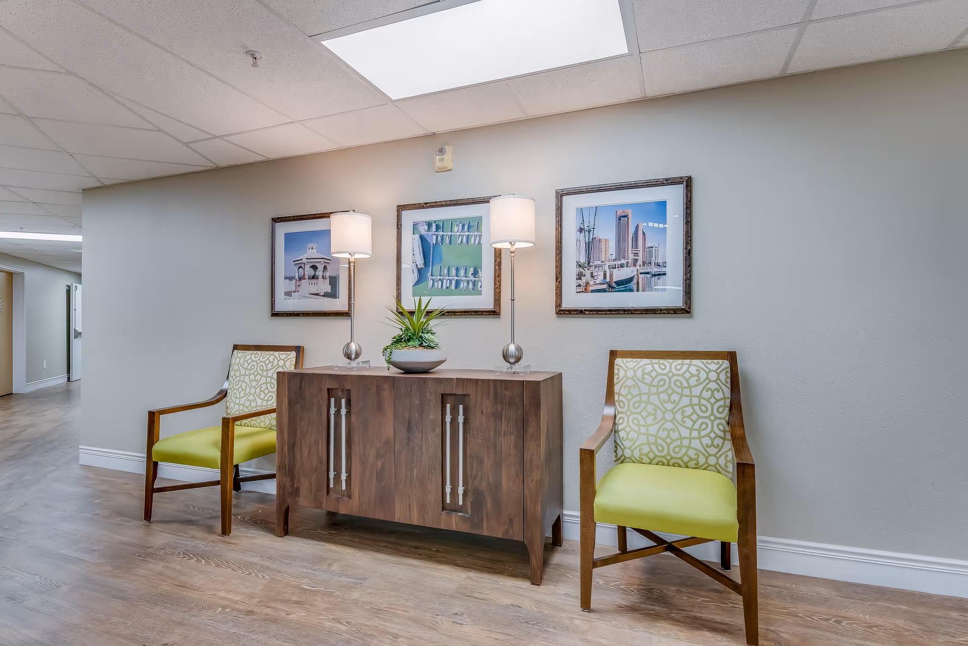 A hallway seating area with two green-upholstered chairs flanking a wooden console table topped with lamps, a plant, and framed artwork on the wall.