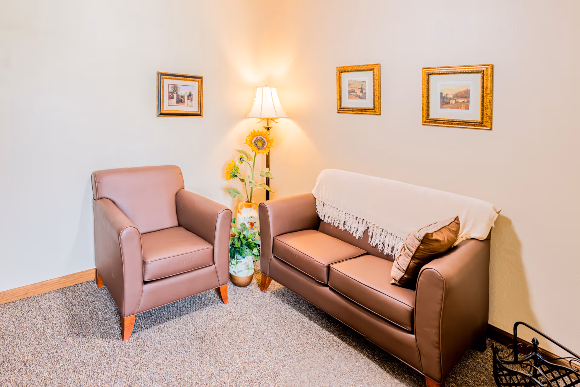 A cozy corner of a living room featuring a brown leather armchair and a matching three-seat sofa with a beige throw draped over the back. A floor lamp with a white shade stands between the two seats, next to a vase with sunflowers and green plants. Three framed pictures hang on the beige wall above the sofa and armchair. The floor is carpeted in a neutral tone.