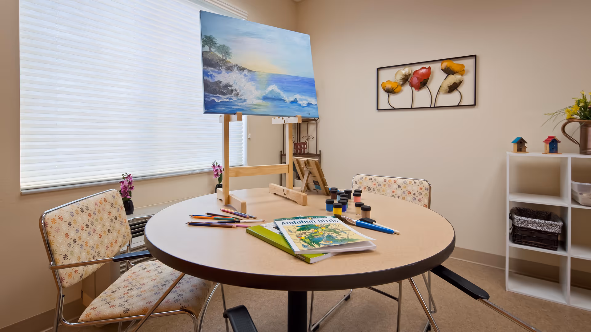 A small art activity room with a round table holding an easel with a painting of a seascape, paint bottles, colored pencils, and coloring books. Two floral-patterned chairs are positioned around the table. A window with blinds and a wall with decorative artwork and a shelving unit with small items are visible in the background.