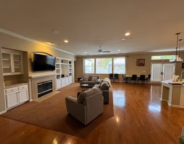 A spacious living room area with hardwood floors and a large brown carpet. There are two gray sofas and a matching armchair arranged around a small round coffee table. A flat-screen TV is mounted above a fireplace with built-in white shelving units on either side. Large windows let in natural light, and a dining table with chairs is visible near the back wall next to glass doors. The room has recessed lighting and a ceiling fan.