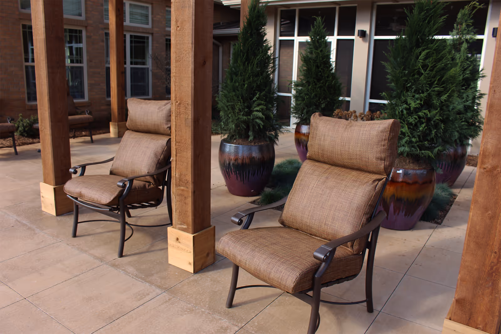 Outdoor patio area with cushioned chairs and large potted evergreen plants, supported by wooden beams, in front of a building with multiple windows.