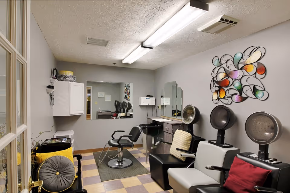 Interior view of a small hair salon or beauty room with two salon chairs, hair dryers, a large mirror on the wall, and colorful wall art. The room has a checkered floor and overhead fluorescent lighting.
