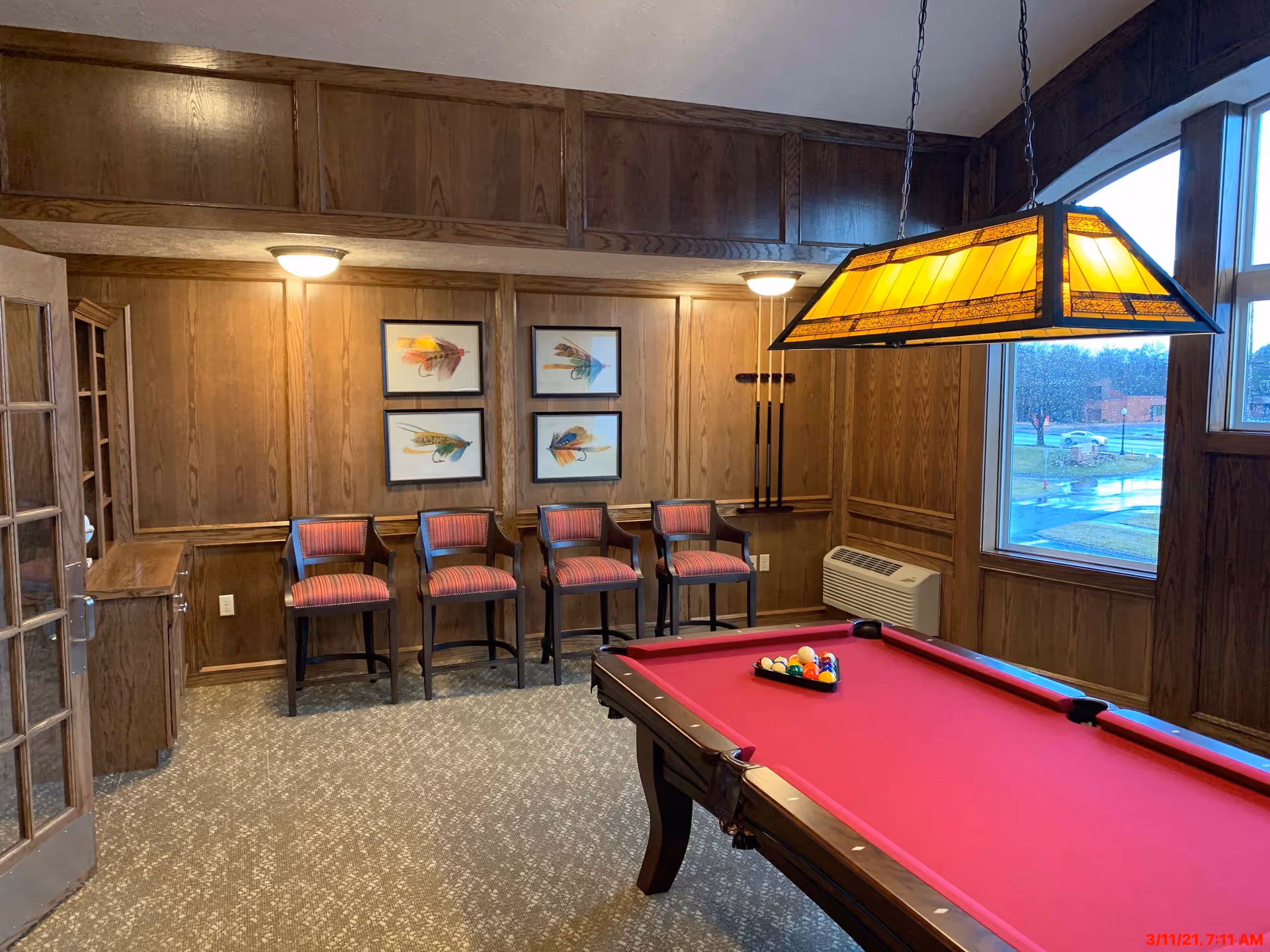Interior room with a red pool table in the foreground, four wooden chairs with red striped cushions lined up against a wood-paneled wall, four framed pictures of colorful feathers on the wall, a hanging yellow stained-glass light fixture above the pool table, a window showing an outdoor view, and a carpeted floor.