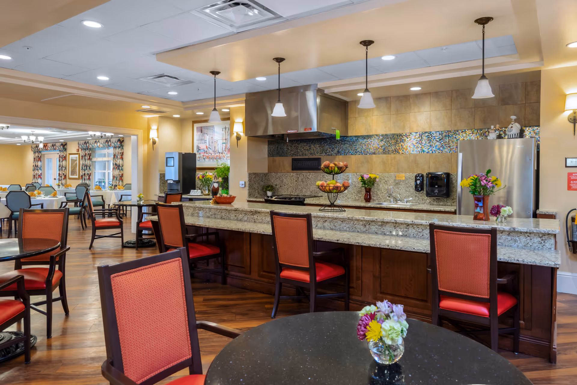 A spacious and well-lit dining area with a kitchen counter and bar stools. The kitchen features a granite countertop, stainless steel refrigerator, stove with a range hood, and decorative tile backsplash. Several tables and chairs with red cushions are arranged around the room, and small flower arrangements are placed on the tables and counter. The background shows more dining tables and windows with patterned curtains.