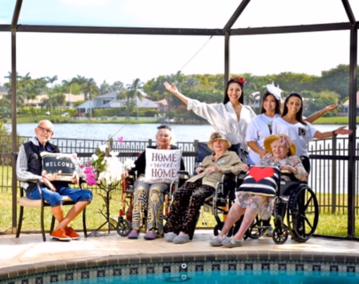 A group of elderly individuals and caregivers posing happily outdoors near a pool with a lake and houses in the background. Three elderly people are seated, two in wheelchairs and one on a chair, holding decorative pillows with welcoming messages. Three caregivers stand behind them, smiling and gesturing with open arms.