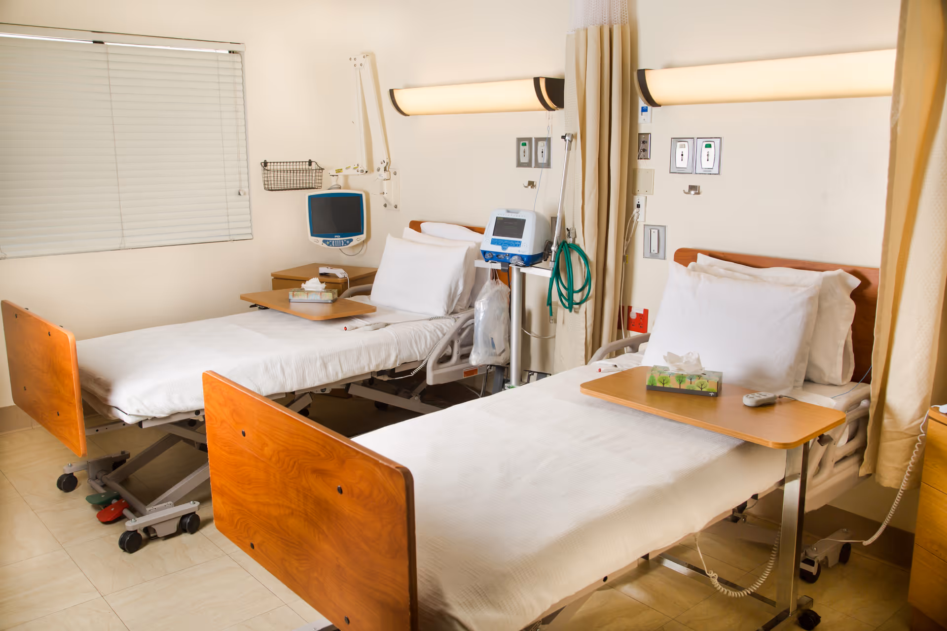 A hospital or healthcare facility room with two adjustable beds, each equipped with white linens, pillows, and overbed tables. Medical monitors and equipment are mounted on the wall behind the beds, and privacy curtains hang between the beds. The room has a window with closed blinds and a clean, clinical appearance.