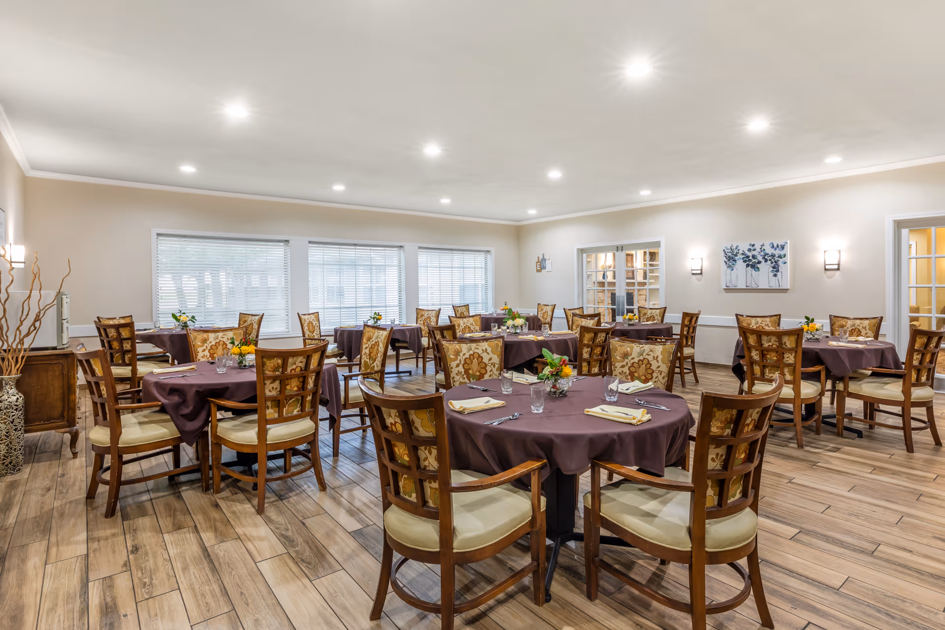 A dining room with multiple round tables covered in dark purple tablecloths, each set with napkins, glasses, and silverware. The chairs have wooden frames with floral patterned cushions. The room has wood flooring, beige walls, recessed ceiling lights, and large windows with blinds. There are decorative elements including a vase with branches and framed artwork on the walls.