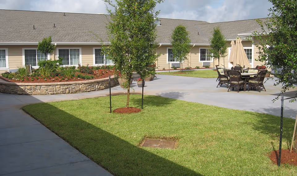 Outdoor courtyard area of a senior living facility with a well-maintained lawn, small trees, a raised flower bed with plants and flowers, and a patio area with a table, chairs, and closed umbrellas. The building with beige siding and multiple windows surrounds the courtyard.