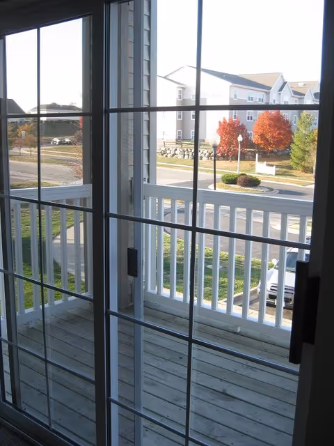 View through a glass sliding door onto a wooden balcony with white railings. Outside, there is a parking lot, a road, and a landscaped area with trees showing autumn foliage and a multi-story residential building in the background.