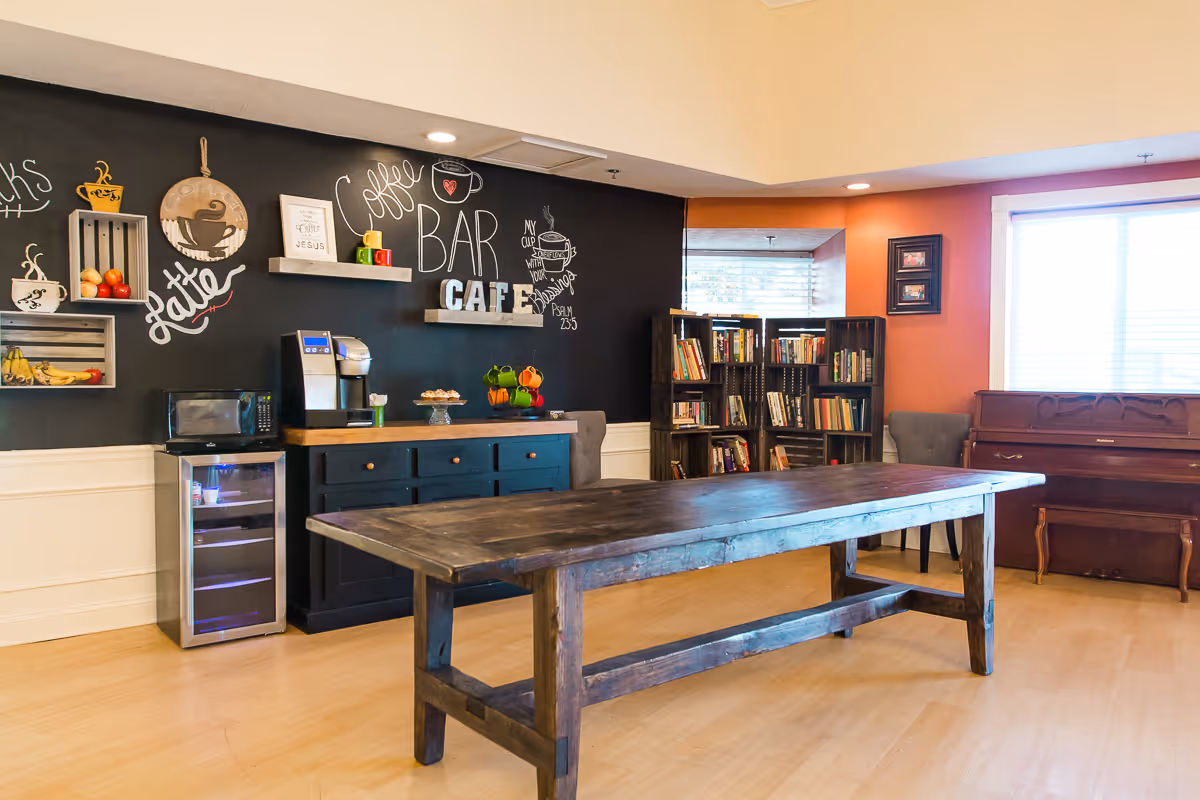 A cozy room with a large wooden table in the center, a black chalkboard wall decorated with coffee-themed drawings and words like 'Coffee Bar Cafe', a small refrigerator, a coffee machine, shelves with fruit and mugs, a bookshelf filled with books, a piano, and two gray chairs near a window with blinds.