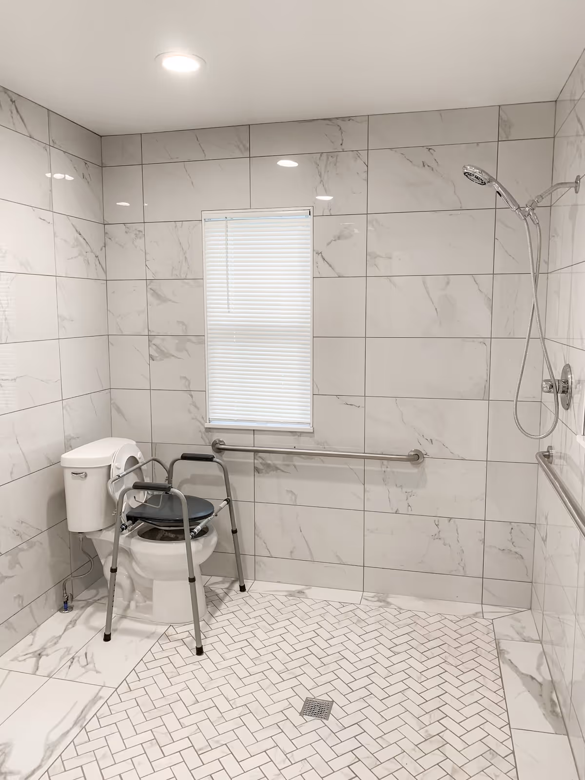 A bathroom with white marble-patterned tiles on the walls and floor. There is a toilet with a raised seat and armrests for accessibility. A handheld showerhead is mounted on the wall to the right, and grab bars are installed on the walls near the toilet and shower area. A small window with closed blinds is centered on the back wall.