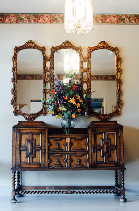 Ornate wooden sideboard topped with a vase of flowers beneath three gilded mirrors and a hanging chandelier.