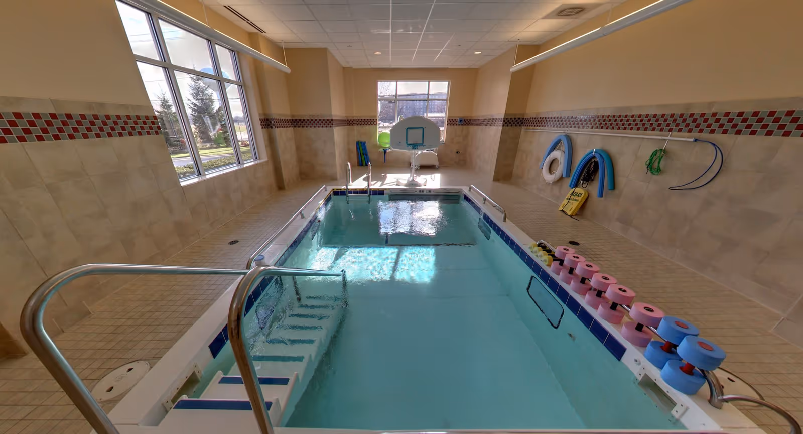 Small indoor therapy pool in a tiled room with handrails, a pool lift, flotation noodles and large windows.