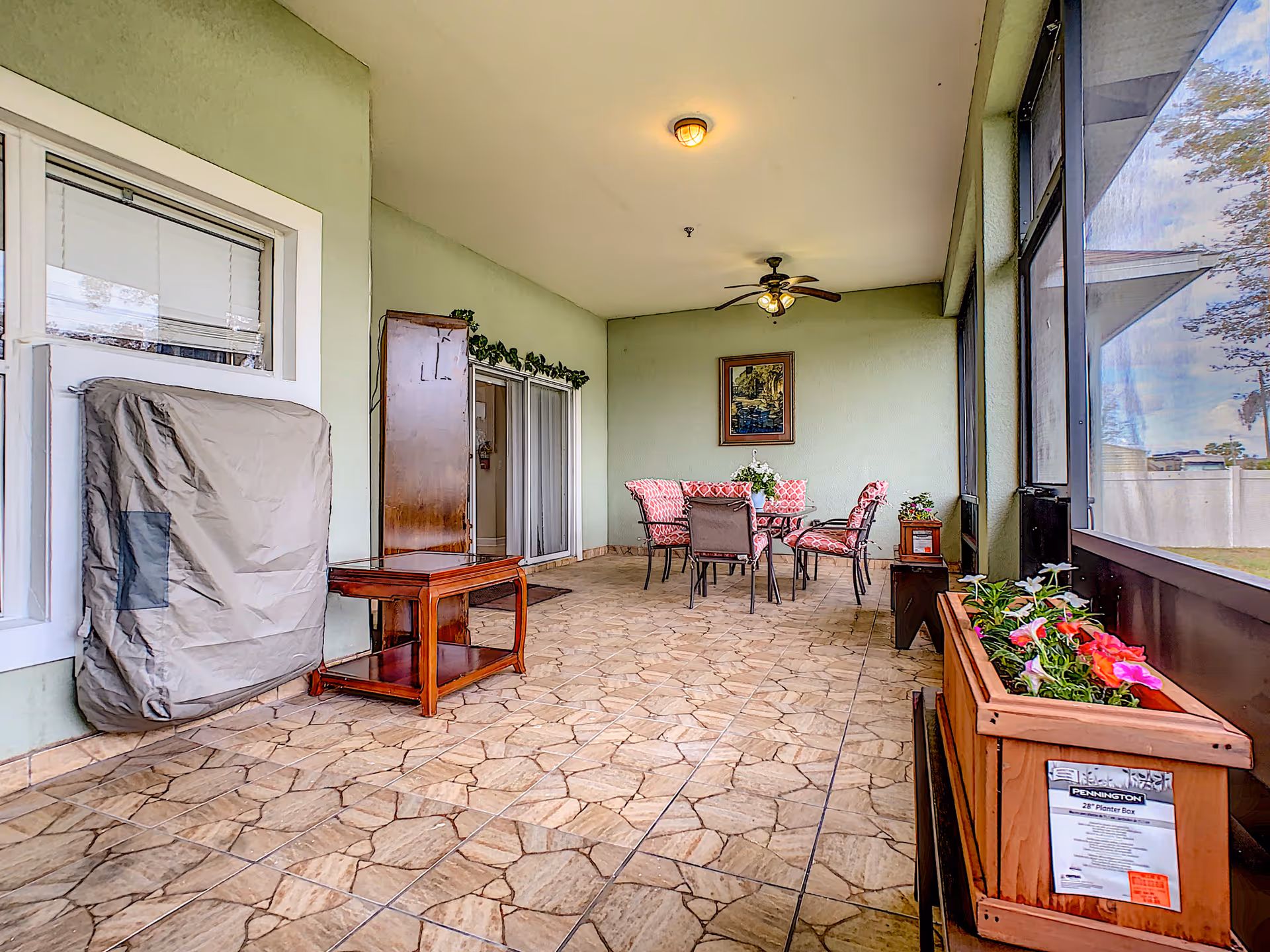 A covered patio area with tiled flooring and green walls. There is a table with four chairs featuring red patterned cushions, a ceiling fan with lights, a framed painting on the wall, and a wooden planter box with flowers on the right side. A covered object and a small wooden table are on the left side near a window and sliding glass doors.