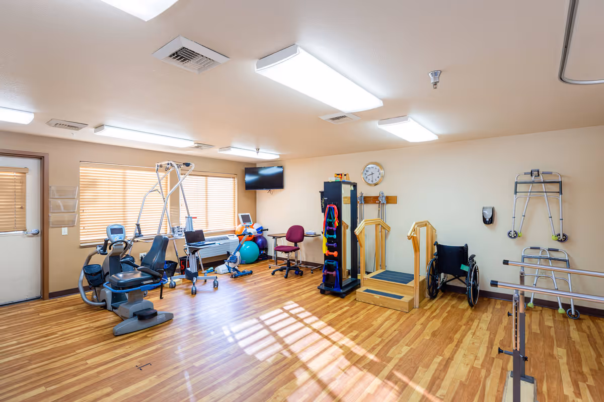 A well-lit physical therapy or rehabilitation room with exercise equipment including a recumbent bike, parallel bars, a wheelchair, walkers, exercise balls, and a rack of colorful dumbbells. The room has wooden flooring, beige walls, a wall clock, and a mounted TV. Large windows with blinds allow natural light to enter.