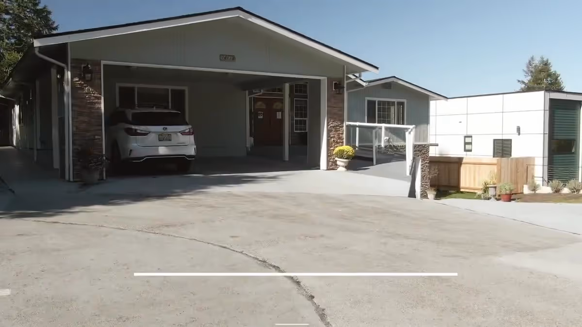 Exterior view of a single-story residential building with a covered carport containing a white SUV. The building has a light gray facade with stone accents and a ramp leading to the entrance. There is a clear blue sky in the background.