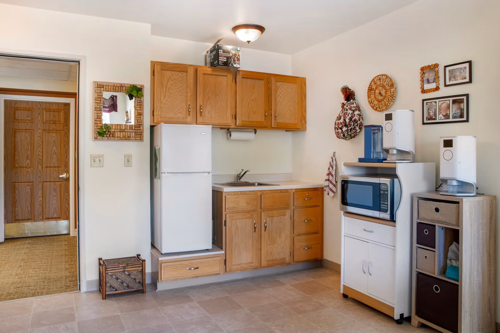 A small kitchen area with wooden cabinets, a white refrigerator, a sink, a microwave on a white stand, and two water dispensers. There are decorative items on the wall including framed photos and a woven wall hanging. A doorway with a wooden door is visible to the left.