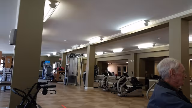Interior view of a rehabilitation and nursing center gym area with exercise equipment including stationary bikes and weight machines. An elderly man is seated on the right side of the image. The room has beige walls, tiled flooring, and ceiling lights.