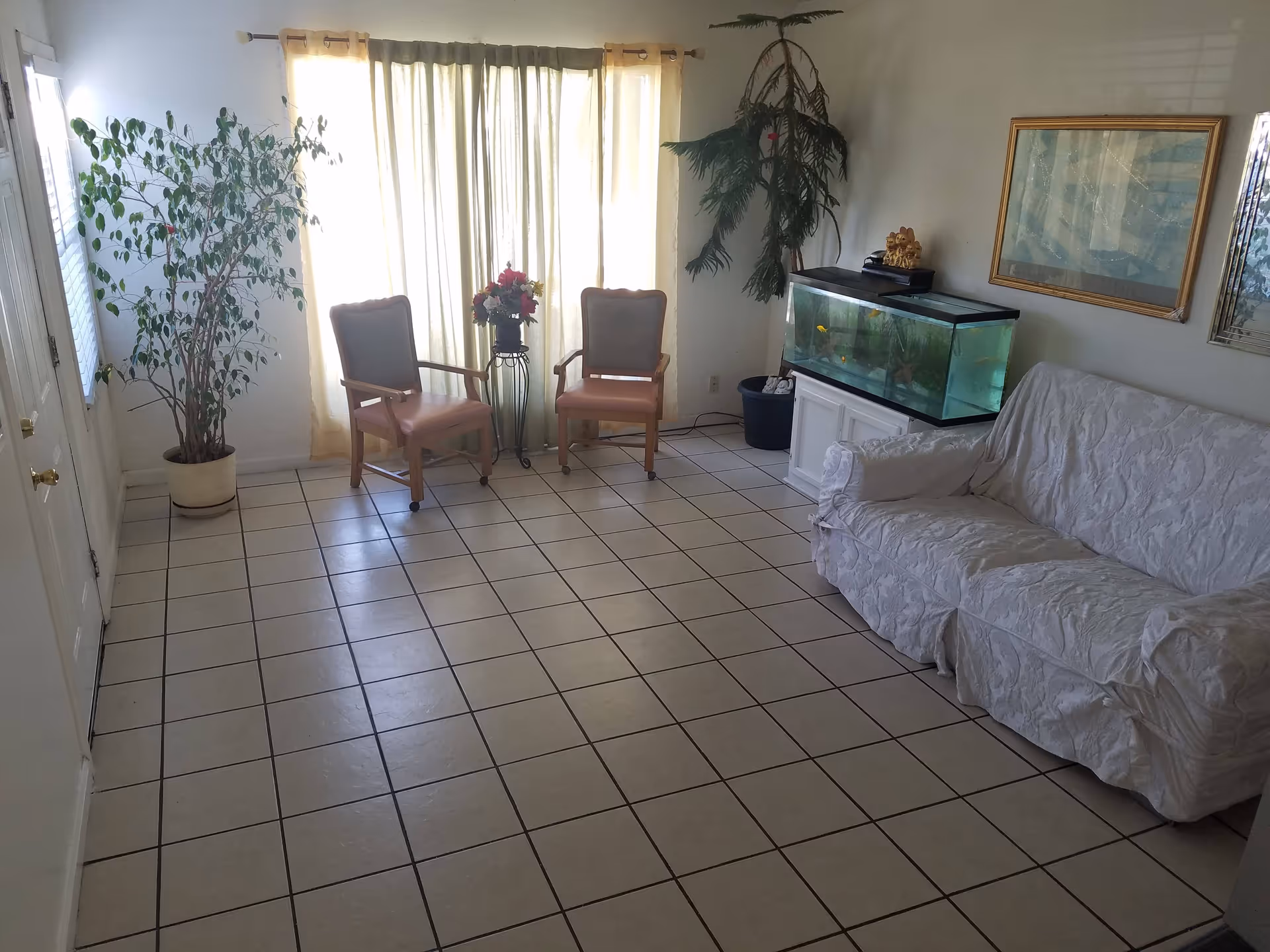 Tiled living room with a covered sofa, two armchairs by a curtained window, an aquarium and potted plants.