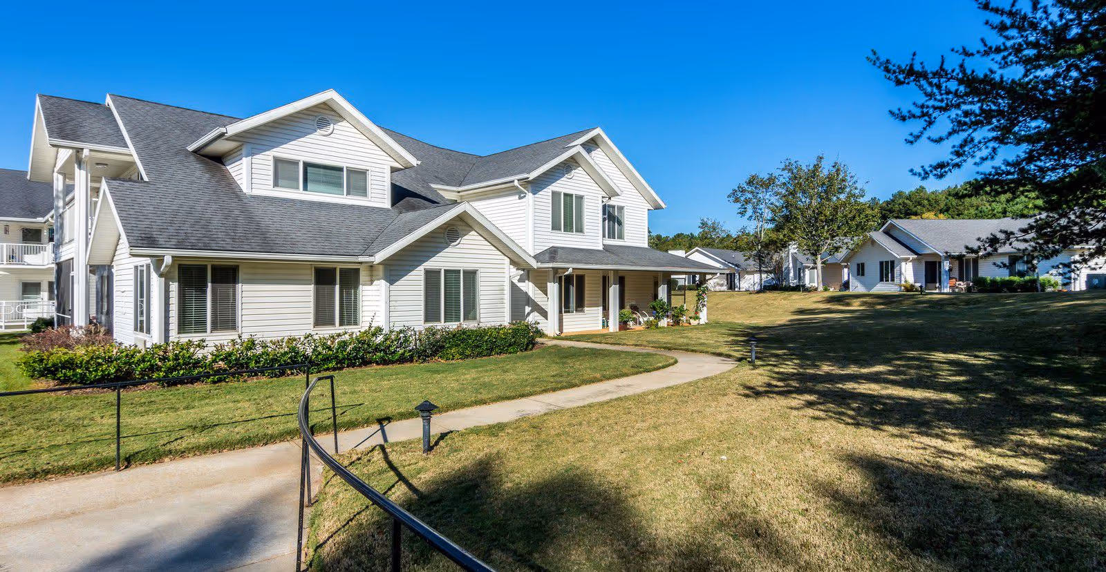 Front exterior of a white two-story residential building on a landscaped senior living campus with a walkway and lawn.