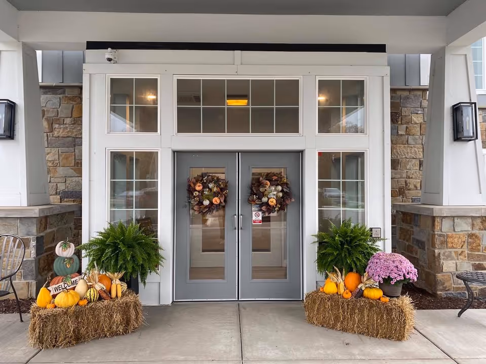 Covered front entrance with double glass doors decorated with fall wreaths, pumpkins, hay bales and potted plants.