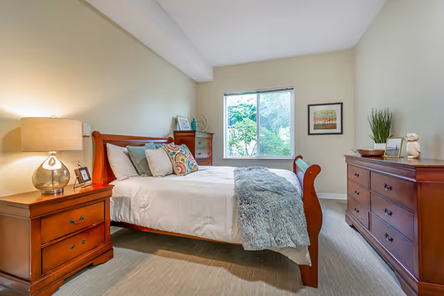 A well-lit bedroom with a wooden bed frame, white bedding, and decorative pillows. There is a wooden nightstand with a lamp and picture frame on the left side of the bed. A wooden dresser with decorative items and a framed picture hangs on the wall to the right. A window with a view of greenery outside is centered on the back wall.