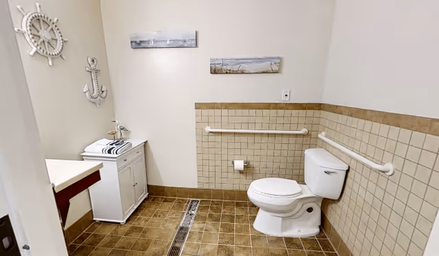 A clean and spacious bathroom with a white toilet, beige tiled walls and floor, white grab bars on the walls, a small white cabinet with folded towels, and nautical-themed wall decorations including a ship wheel and an anchor.