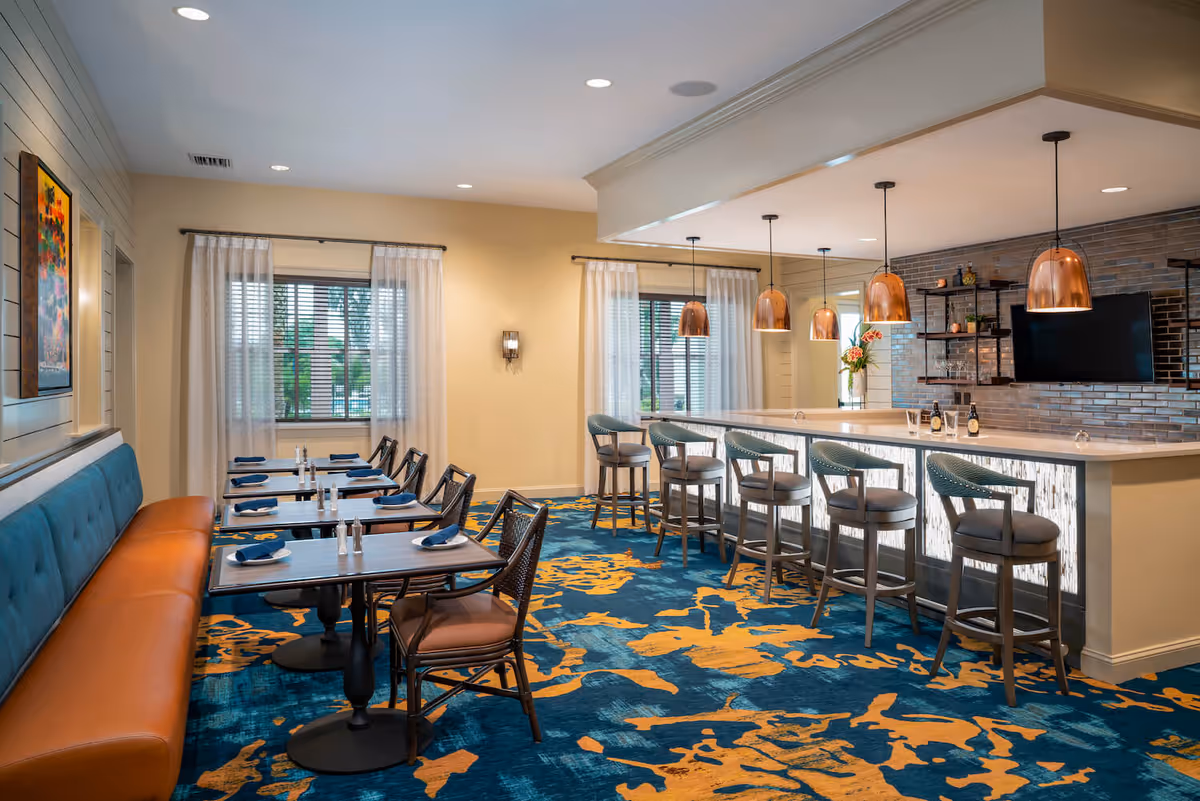 A bright and modern dining area featuring a long bar with six high chairs and copper pendant lights hanging above. To the left, there are several small dining tables with chairs and a long cushioned bench along the wall. The room has large windows with sheer white curtains, a colorful blue and orange patterned carpet, and a wall-mounted TV above the bar area.