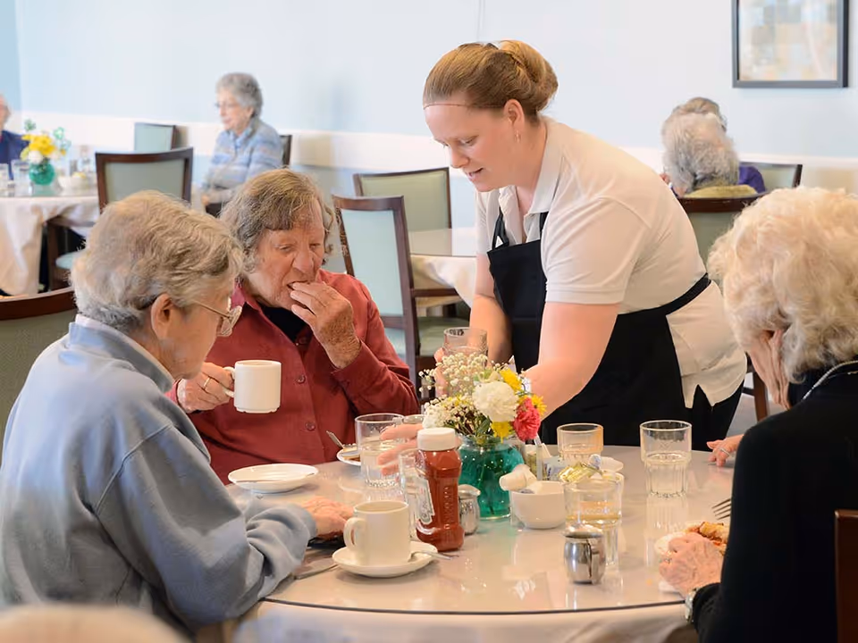A dining room scene in a senior living facility where a caregiver in a white shirt and black apron is serving drinks to elderly residents seated around a table with cups, glasses, and a flower vase.