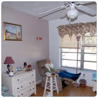A senior woman sitting in a recliner chair in a cozy room with a ceiling fan, a window with floral valance curtains, a white dresser with a lamp and decorative items, and a small stool in front of the chair.