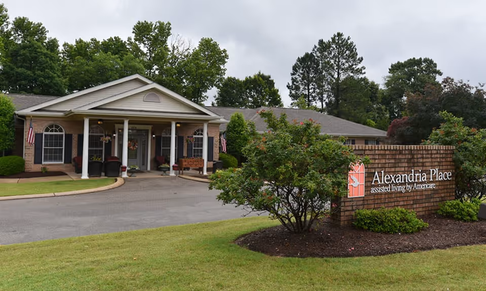 Front entrance of Alexandria Place assisted living facility with a porte-cochere, columns, driveway, and a brick sign in the landscaped foreground.