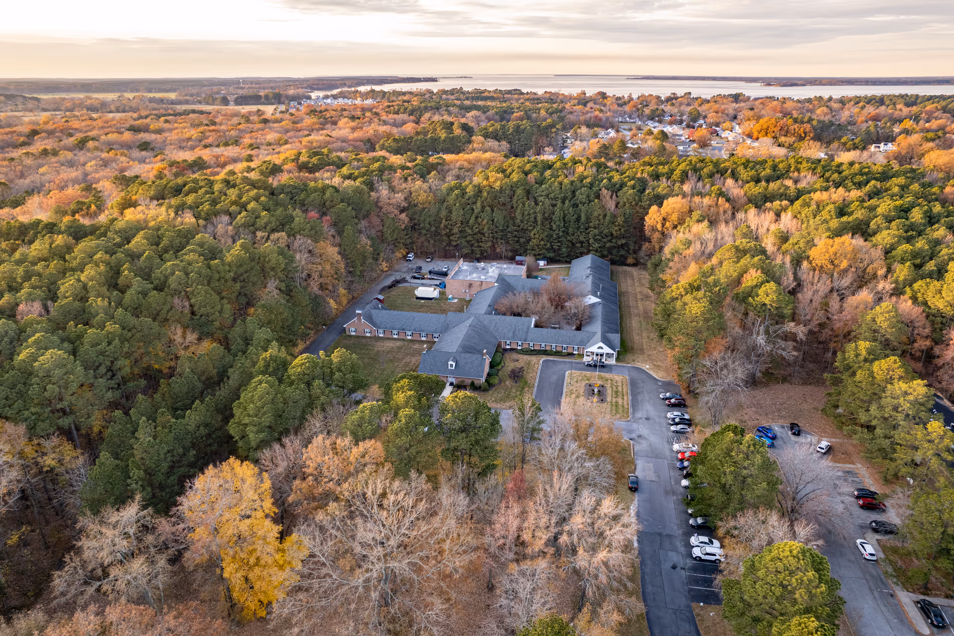 Aerial view of Autumn Lake Healthcare at Chesapeake Woods surrounded by dense trees with autumn foliage. The building is a single-story structure with a gray roof and a parking lot with several cars. The surrounding area is heavily wooded with a mix of green and orange-brown trees under a cloudy sky.