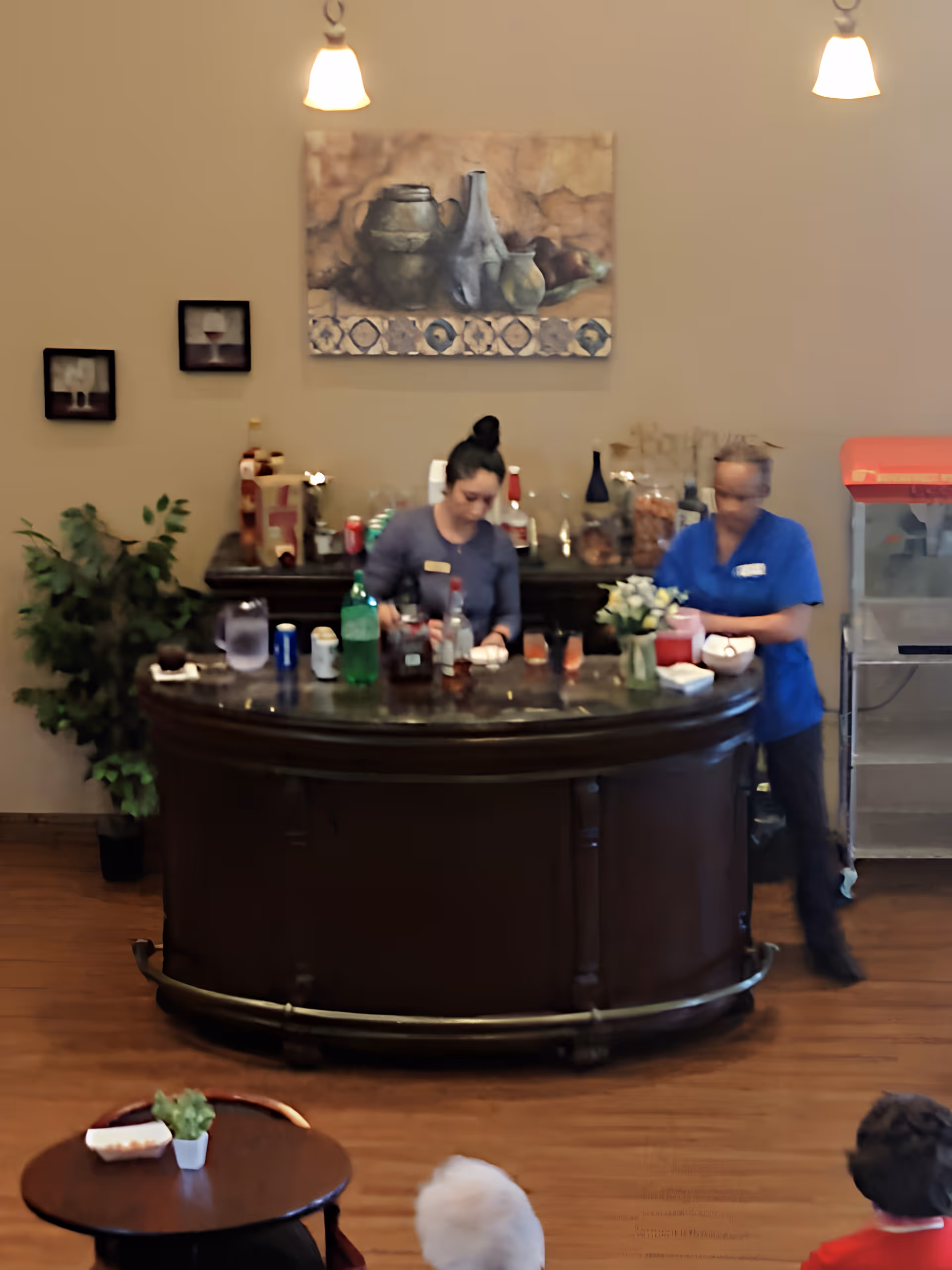 Two staff members working behind a curved service bar in a communal dining/lounge area with a few residents visible.