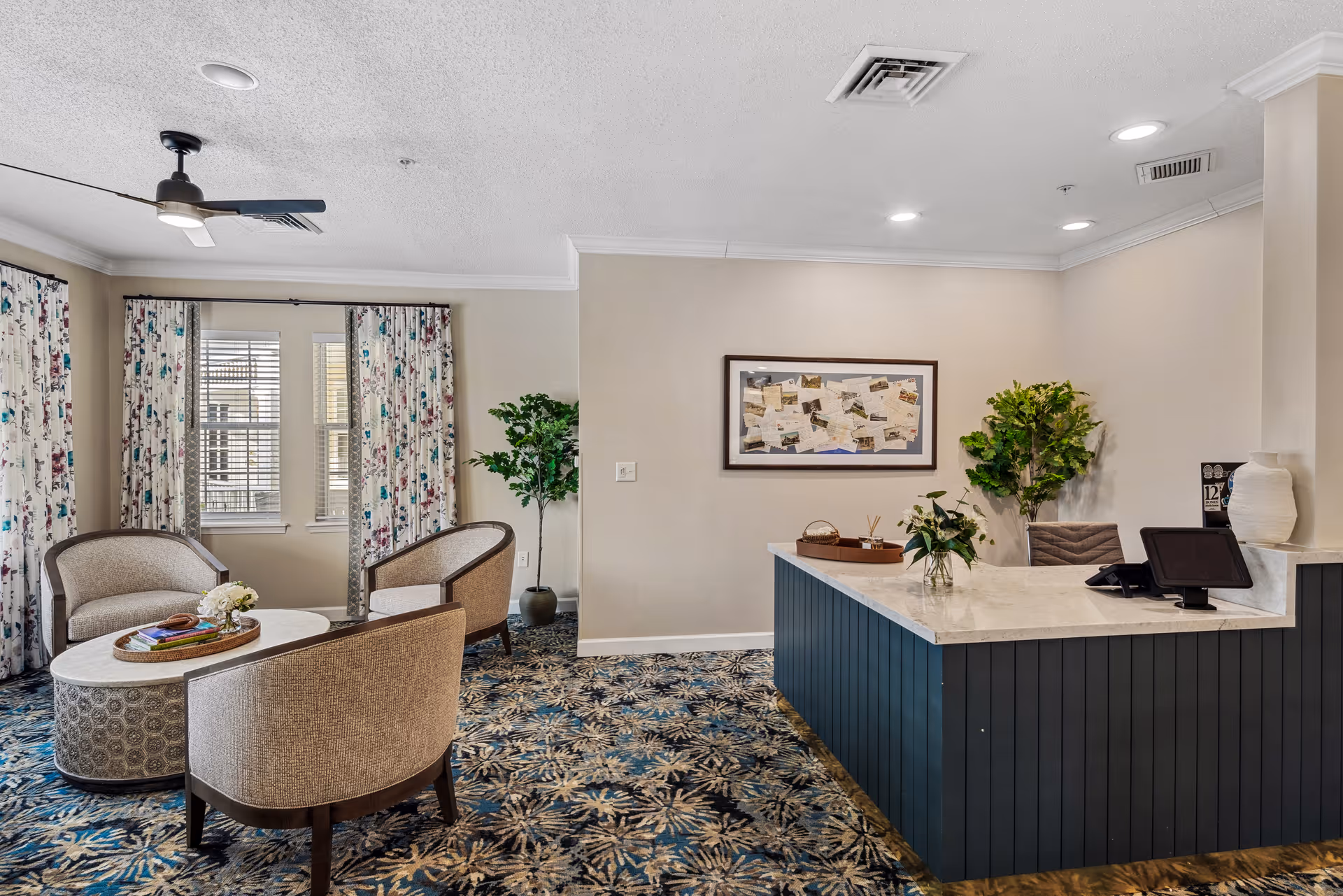 Reception area with a marble countertop desk, a computer monitor, and decorative plants. To the left, there is a seating area with three upholstered chairs around a round coffee table. The room has patterned carpet, floral curtains on windows, and a ceiling fan.