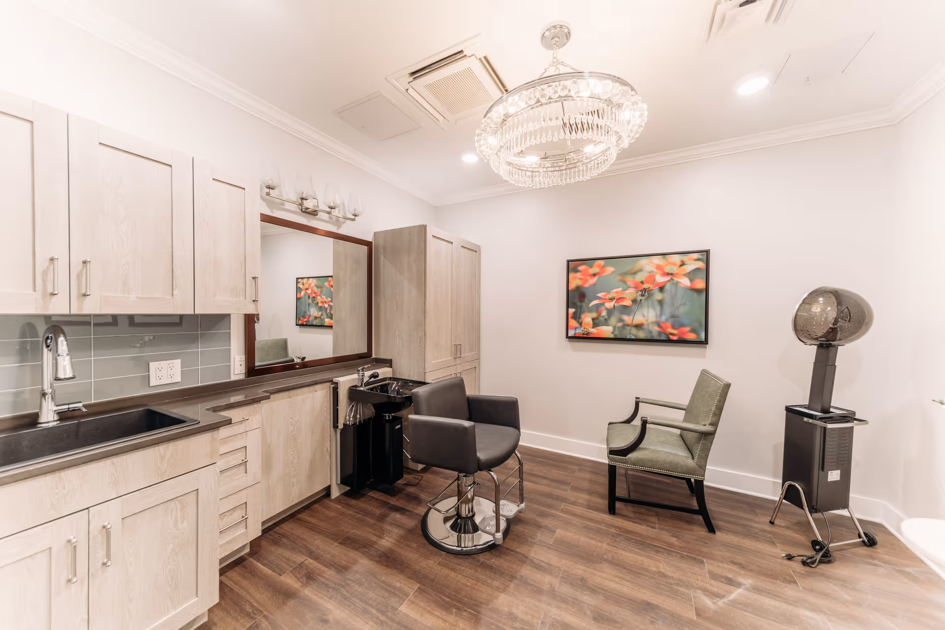 A modern salon room with light wood cabinetry, a black salon chair in front of a large mirror, a green upholstered chair, a hair dryer on a stand, and a colorful floral painting on the wall. The room has wood flooring and a decorative chandelier on the ceiling.