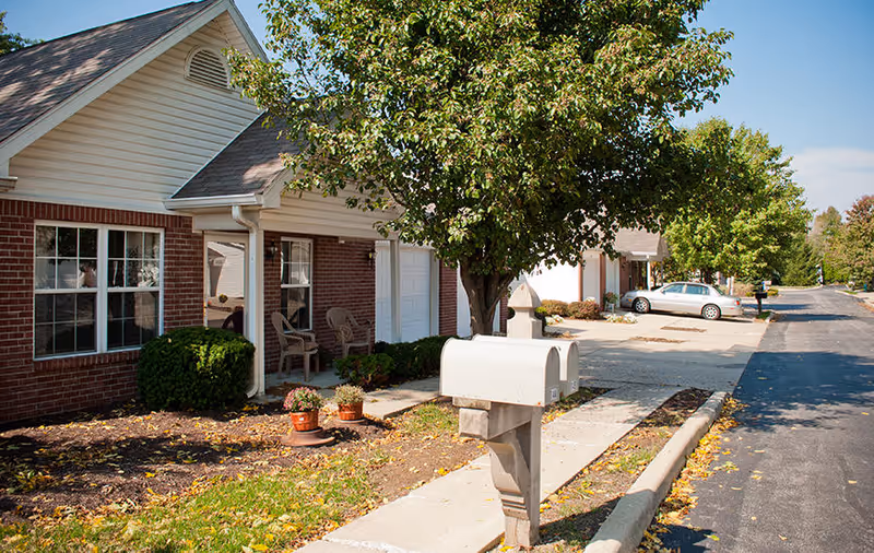 Front of a row of brick-and-siding senior living units with a mailbox, small porches, trees, and parked cars along the driveway.