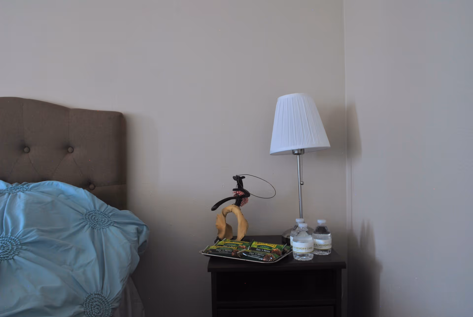 A bedside table next to a bed with a brown tufted headboard and a blue pillow. On the table, there is a silver lamp with a white lampshade, a small decorative item, two bottles of water labeled 'Lotus Villa & Memory Care,' and a tray with packets of Nature Valley granola bars.