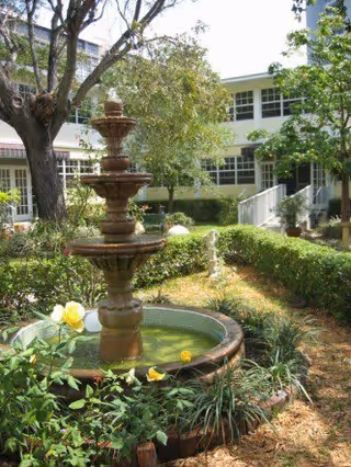 A multi-tiered water fountain in the center of a garden area with green bushes, yellow flowers, and trees. In the background, there is a two-story building with white walls and multiple windows.