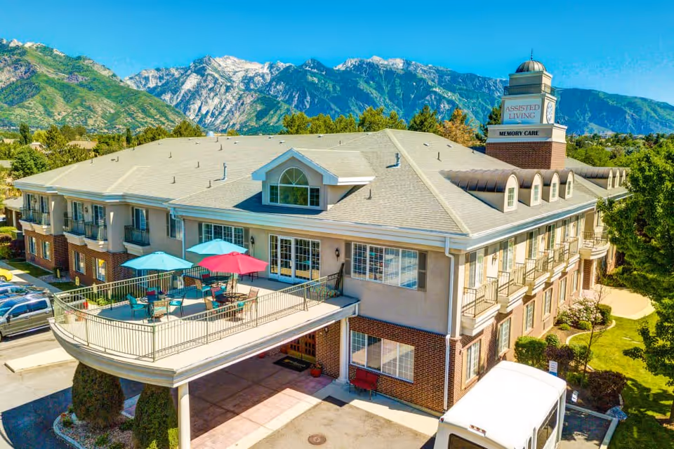 Exterior view of a two-story assisted living facility with a balcony patio featuring tables and colorful umbrellas. The building has a sign indicating 'Assisted Living' and 'Memory Care'. Mountains and clear blue sky are visible in the background.