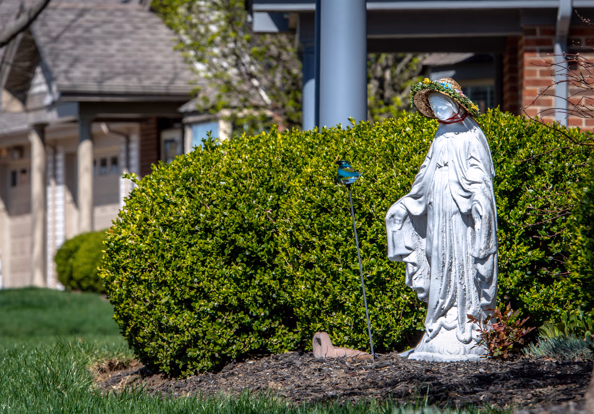 A white statue of a robed figure wearing a straw hat decorated with flowers, standing in a garden bed with mulch and green bushes in the background. A small decorative blue bird is perched on a metal stake next to the statue. Residential buildings with garages are visible in the background.