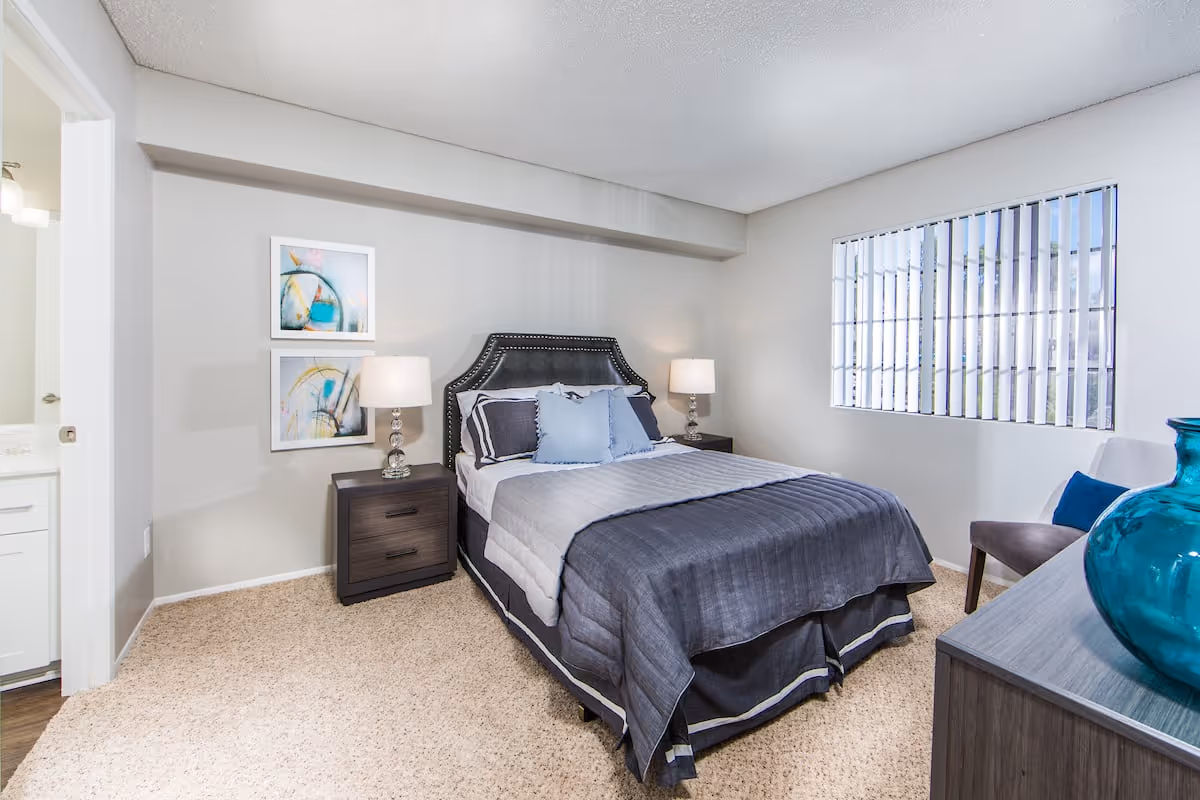 A neatly made bed with a dark upholstered headboard and gray bedding in a bedroom. There are two matching nightstands with lamps on either side of the bed. Two abstract paintings hang on the wall above the left nightstand. A window with vertical blinds is on the right wall, and a gray dresser with a large blue vase is in the foreground. A doorway to a bathroom is visible on the left side of the image.