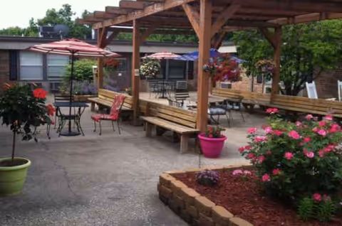 Outdoor courtyard at a senior living facility with pergola, benches, tables, umbrellas, and flowers.