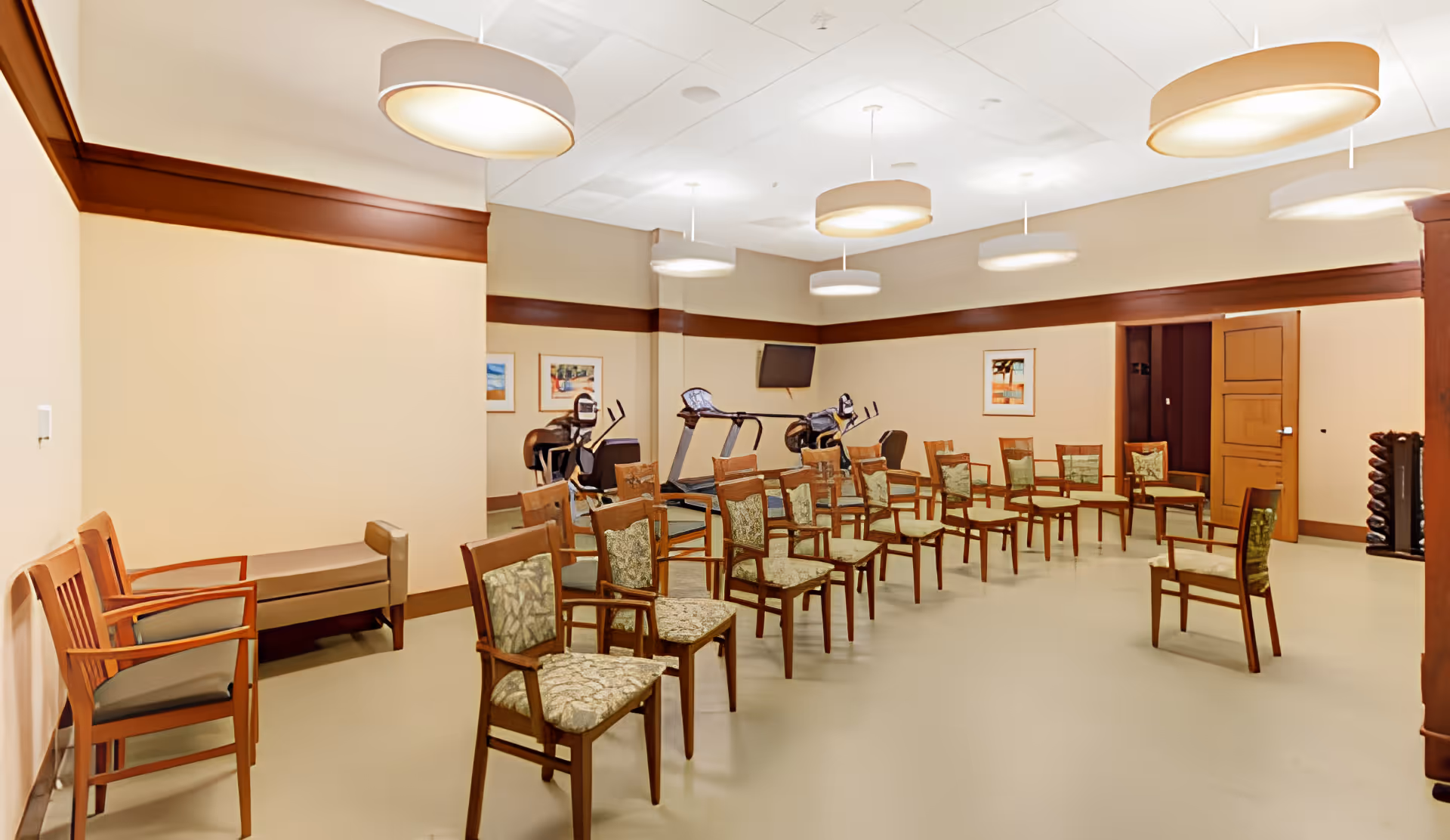 A spacious room with multiple wooden chairs arranged in rows facing a wall-mounted TV. The room has beige walls with dark wood trim and several large round ceiling lights. In the background, there are two exercise bikes and a treadmill, along with framed artwork on the walls. There is also a bench and a few additional chairs along one wall.