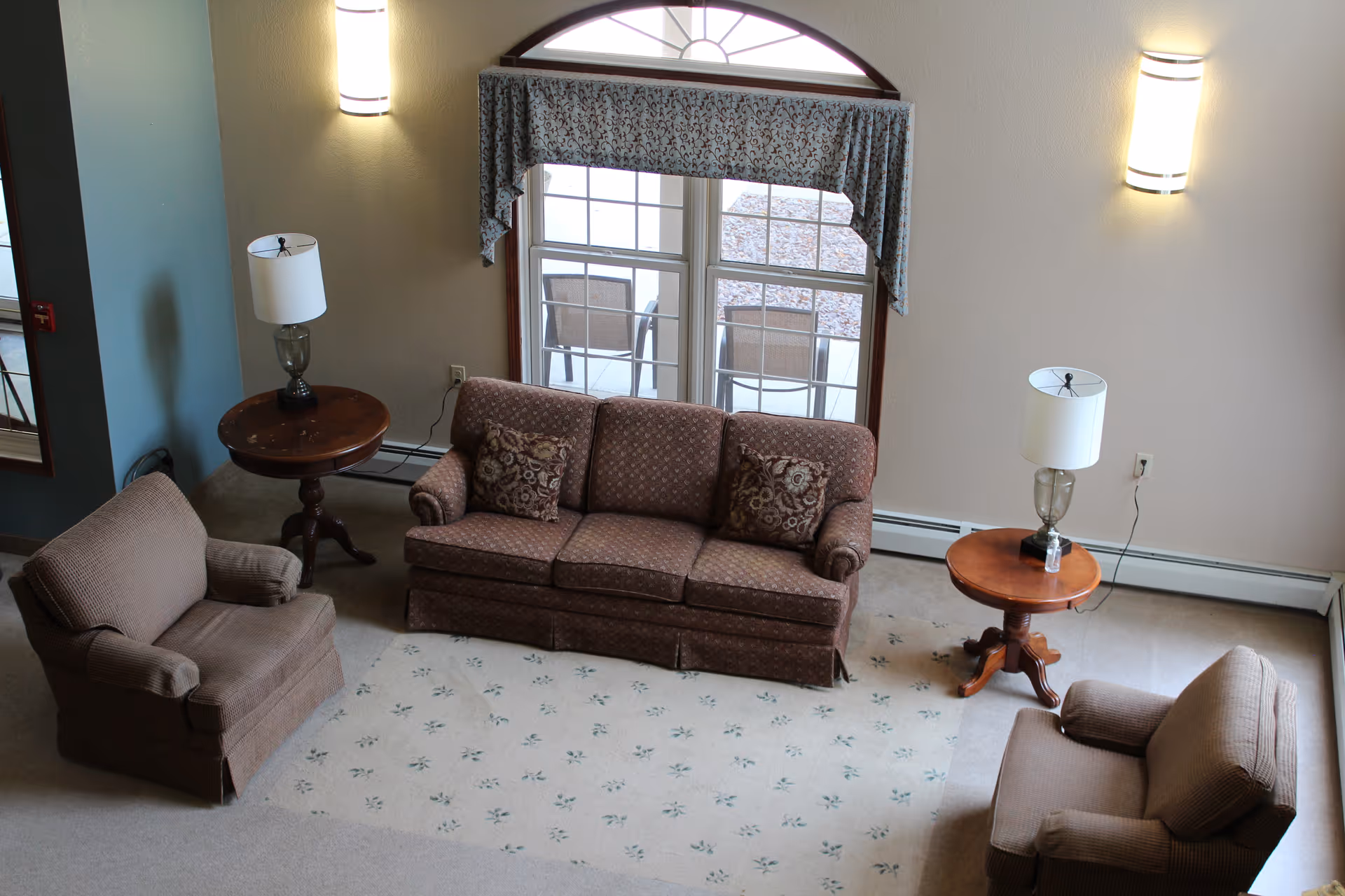 A cozy living room area with a brown patterned sofa and two matching armchairs arranged around a light-colored carpet with a subtle floral pattern. There are two wooden side tables, each with a lamp, positioned on either side of the sofa. The room has a large window with a decorative valance, allowing natural light to enter. The walls are light-colored with two wall-mounted lights.