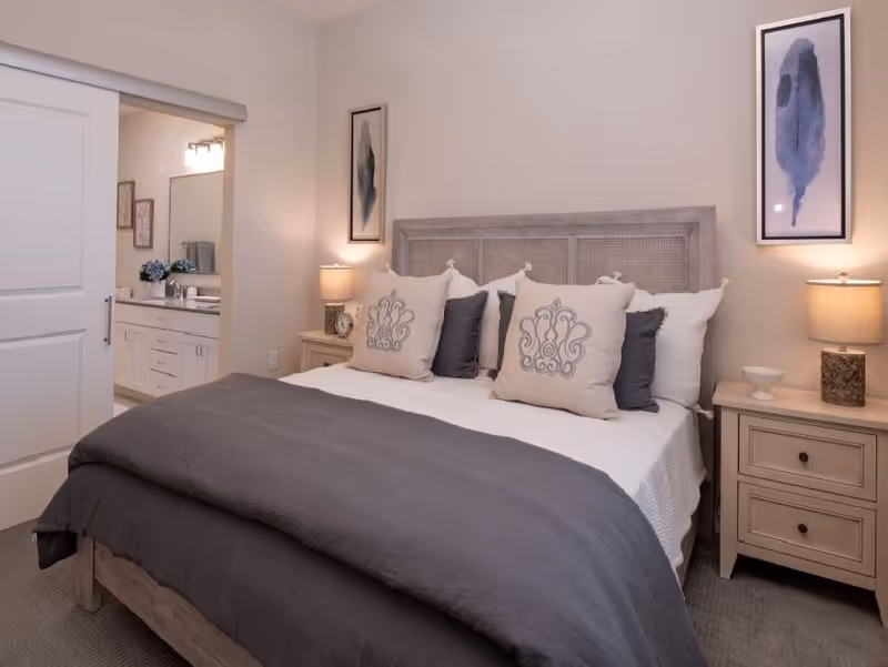 A neatly made bed with a gray comforter and multiple decorative pillows in a bedroom. There are two nightstands on either side of the bed, each with a lamp. Two framed abstract artworks hang on the wall above the nightstands. A sliding door on the left side of the image leads to a bathroom with a vanity and mirror visible.