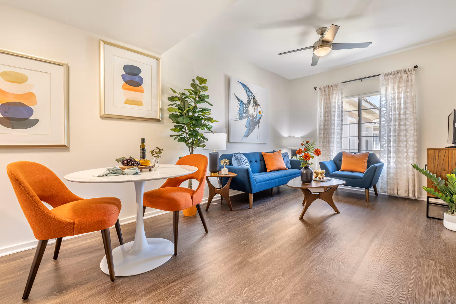 A bright and modern living room with a blue sofa and matching armchair, both adorned with orange pillows. There is a wooden coffee table with a vase of orange flowers, a side table with a lamp, and a ceiling fan above. Two orange chairs are positioned around a small white round dining table with a bottle, grapes, and a napkin on it. The room has light-colored walls with abstract artwork and a large window with patterned curtains letting in natural light. The floor is wood-style laminate.