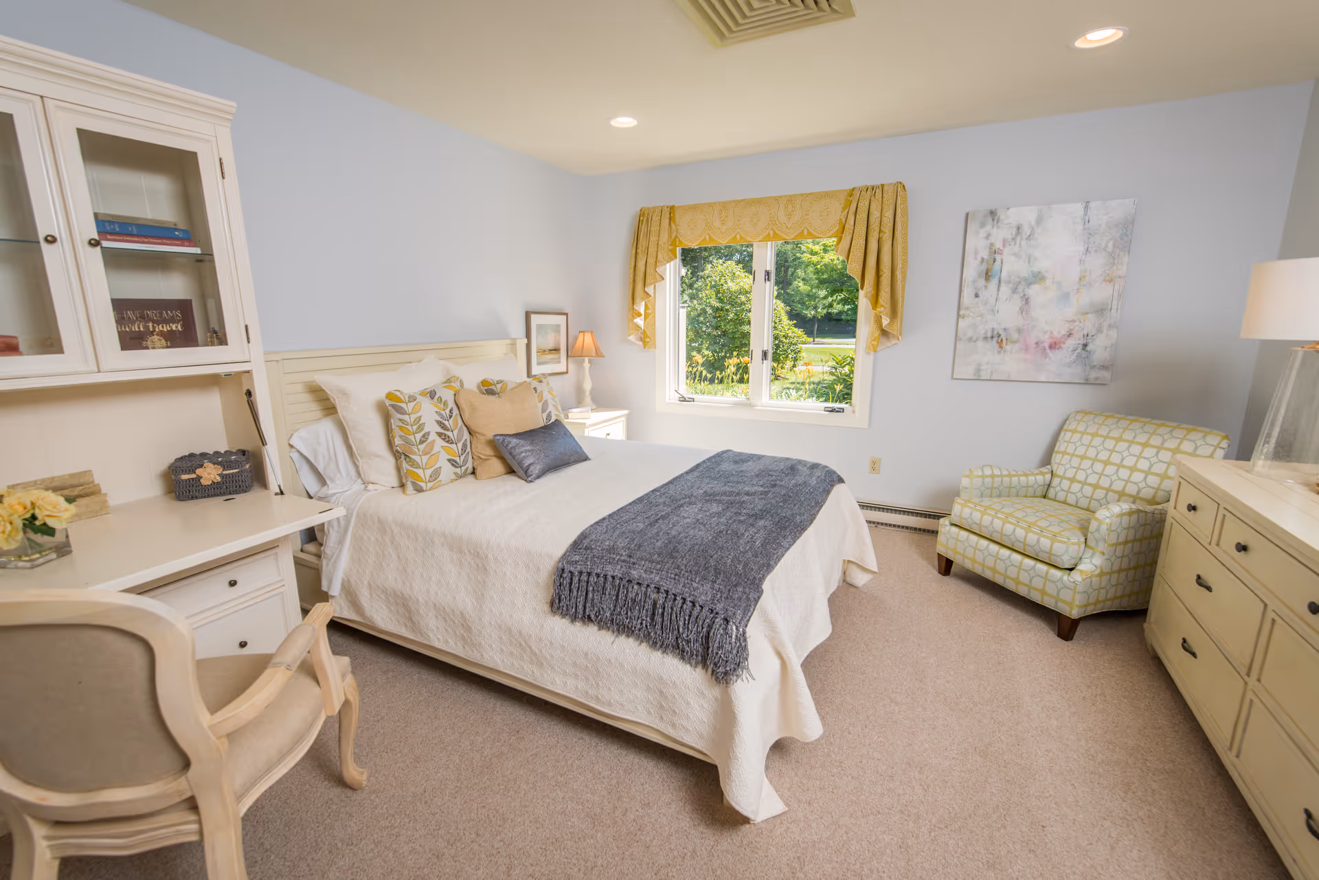 A bright and cozy bedroom with a neatly made bed featuring multiple decorative pillows and a gray throw blanket. The room includes a white desk with a chair, a white dresser with a lamp, a patterned armchair, a window with yellow curtains, and light blue walls adorned with artwork.