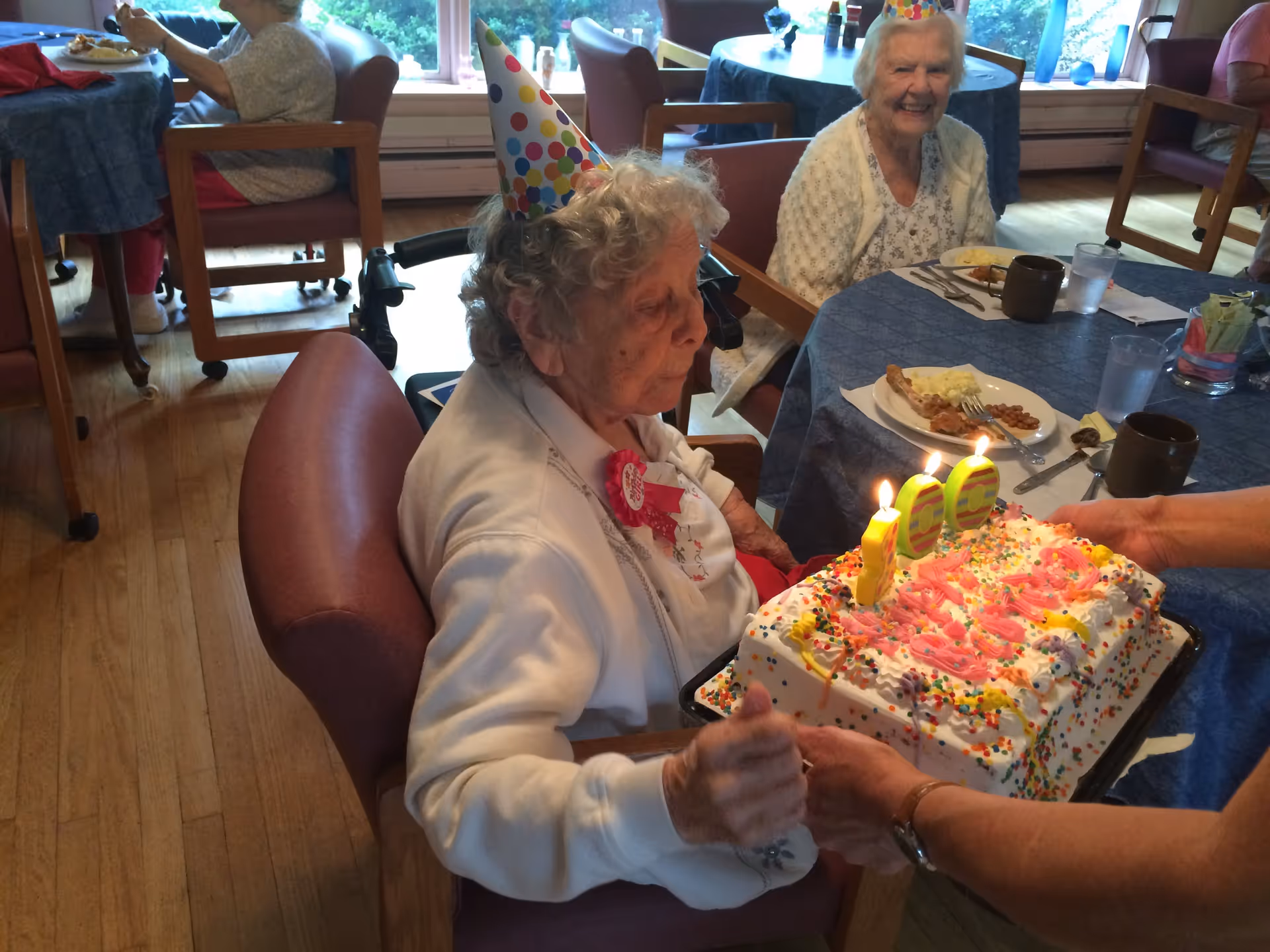 An elderly woman in a party hat sits at a dining table while a decorated birthday cake with lit candles is presented to her.